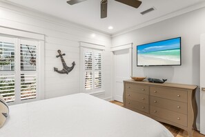 A bedroom with a bed, a dresser, a television, and a window with shutters. - Discover Southern Tides with Bonheur Vacations! (Watersound)