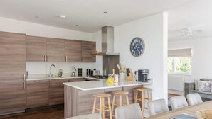 A modern kitchen with wooden cabinets, a central island with stools, a coffee machine, and a clock on the wall. - Lakeside Villa w/ Pool & Tennis in Cotswolds (Gloucestershire)
