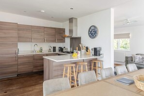 A modern kitchen with wooden cabinets, a central island with stools, a coffee machine, and a clock on the wall. - Lakeside Villa w/ Pool & Tennis in Cotswolds (Gloucestershire)
