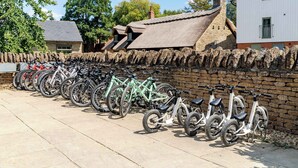 A row of bicycles parked against a stone wall, with a building and trees in the background. - Lakeside Villa w/ Pool & Tennis in Cotswolds (Gloucestershire)