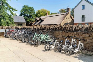 A row of bicycles parked against a stone wall, with a building and trees in the background. - Lakeside Villa w/ Pool & Tennis in Cotswolds (Gloucestershire)