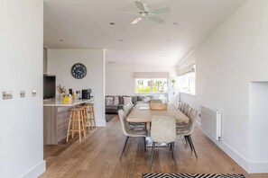 A modern kitchen with a central island, a dining area with a wooden table and chairs, and a living area with a sofa and a large window. - Lakeside Villa w/ Pool & Tennis in Cotswolds (Gloucestershire)