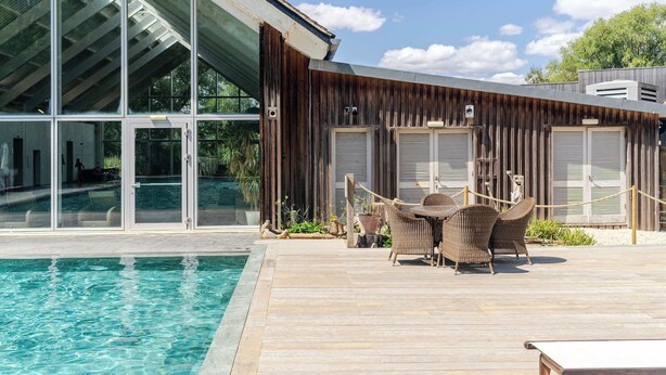 A modern building with large glass windows, a wooden deck, and a pool area with wicker furniture. - Lakeside Villa w/ Pool & Tennis in Cotswolds (Gloucestershire)