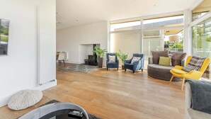 A spacious living room with a large window, a ceiling fan, and a variety of seating options including armchairs and a sofa. - Lakeside Villa w/ Pool & Tennis in Cotswolds (Gloucestershire)