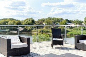 A deck with wicker furniture overlooking a body of water and greenery. - Lakeside Villa w/ Pool & Tennis in Cotswolds (Gloucestershire)