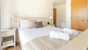 A hotel room with a bed, bedside tables, a lamp, a wooden door, and a wall socket. - Lakeside Villa w/ Pool & Tennis in Cotswolds (Gloucestershire)