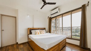 A bedroom with a wooden bed, a ceiling fan, an air conditioning unit, and a large window with curtains. - lavender residense MIDC andheri east (Mumbai)