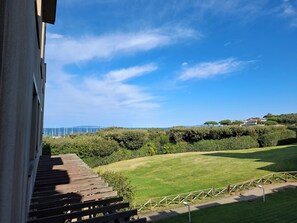 A view of a grassy area with a wooden deck and a fence, overlooking a body of water and distant buildings. - Punta Ala: Studio Apartment with Ocean View - Climate - Parking Space - Beaches (Punta Ala)