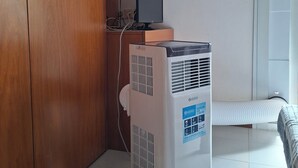 Et værelse med en træskab, en hvid aircondition, en computer på et træskrivebord og et vindue med udsigt til grøn vegetation.