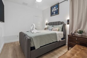 A modern bedroom with a grey bed, a bedside table, a nightstand, a wall-mounted light, and a painting. - The Retreat at Drew | 3 En Suites Beds + Workspace (Houston)