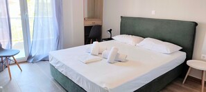 Una cama bien hecha con sábanas y toallas blancas, un cabecero tapizado en verde y una silla junto a la ventana.