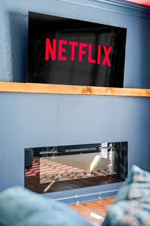 A wall-mounted television displaying the Netflix logo, a lower screen showing a scene from a movie or TV show, and a wooden shelf above the lower screen. - Flames Fan House | *3 MIN to LU* | Sleeps 10! (Lynchburg)
