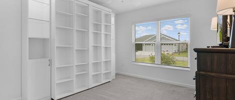 Una habitación con un armario blanco grande, un ventilador de techo, una ventana con vista a una casa y una lámpara sobre un aparador.