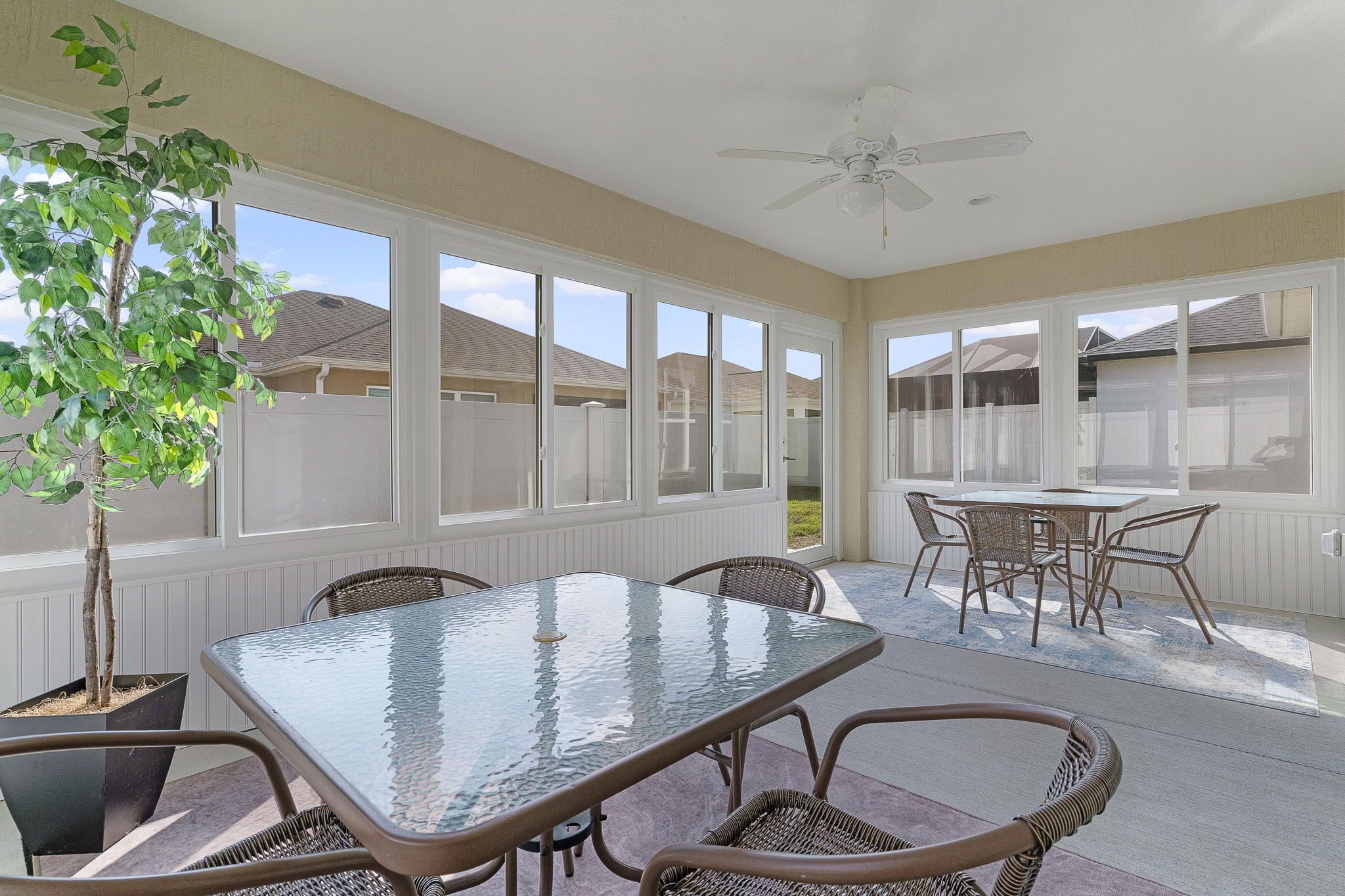 A covered patio with a glass-top table and wicker chairs, surrounded by large windows.