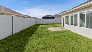 A fenced backyard with a well-maintained lawn, a patio area, and a building with large windows. - Dog-Friendly Citrus Grove Home with Golf Cart, Fenced Yard & Lanai (The Villages)