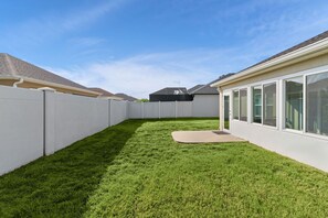 Ein umzäunter Garten mit gepflegtem Rasen, einer Terrassenfläche und einem Gebäude mit großen Fenstern.