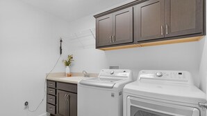 A laundry room with two white washing machines, a dryer, a sink, and a cabinet above the machines. - Dog-Friendly Citrus Grove Home with Golf Cart, Fenced Yard & Lanai (The Villages)