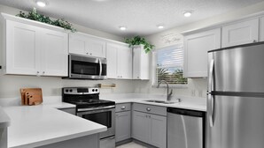 Cocina moderna con electrodomésticos de acero inoxidable, armarios blancos y una ventana con persianas.