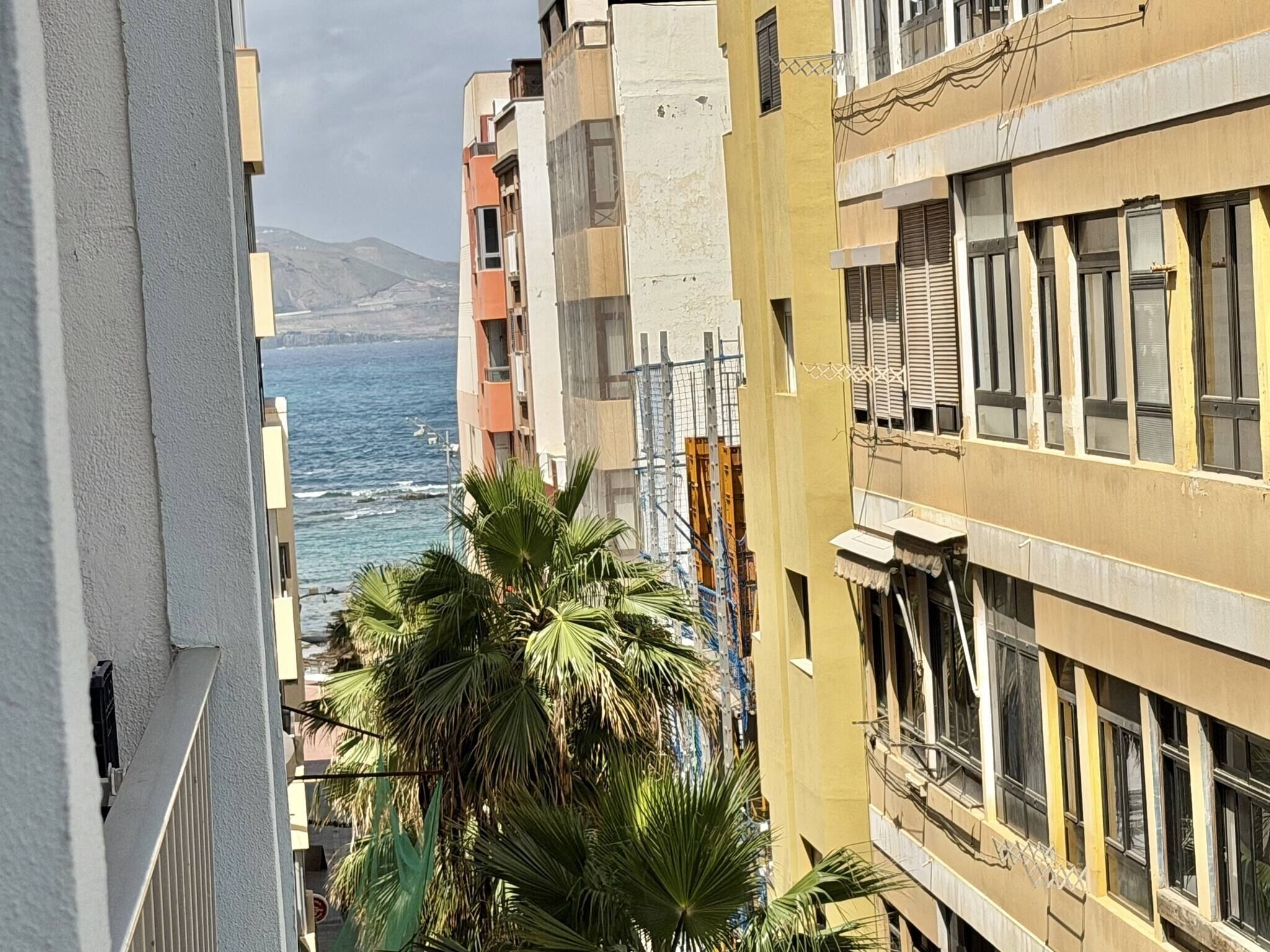 Un edificio costero con balcones que dan al mar, palmeras y montañas al fondo.