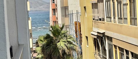 A coastal building with balconies overlooking the sea, palm trees, and a mountainous backdrop.