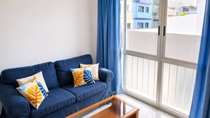 A blue sofa with leaf-patterned pillows, a wooden coffee table, and a view of buildings through a window with blue curtains. - Las Canteras | 1 min to the beach | Ocean view | For 2 guests (Las Palmas de Gran Canaria)