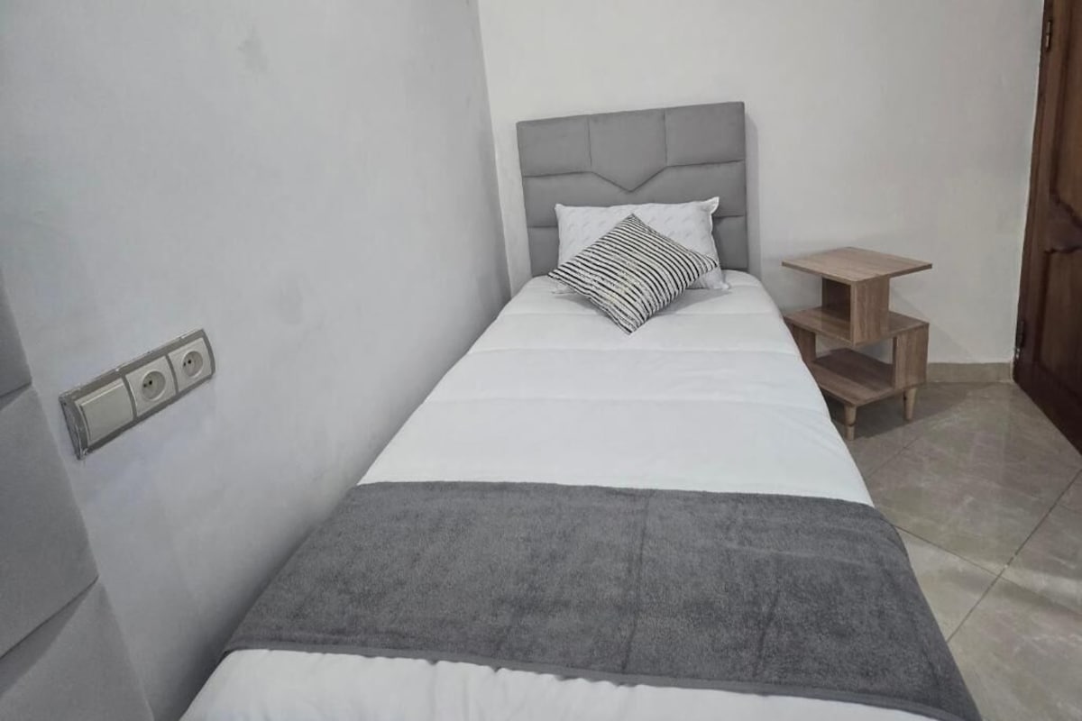 A single bed with a grey headboard, a bedside table, and a power outlet on the wall.
