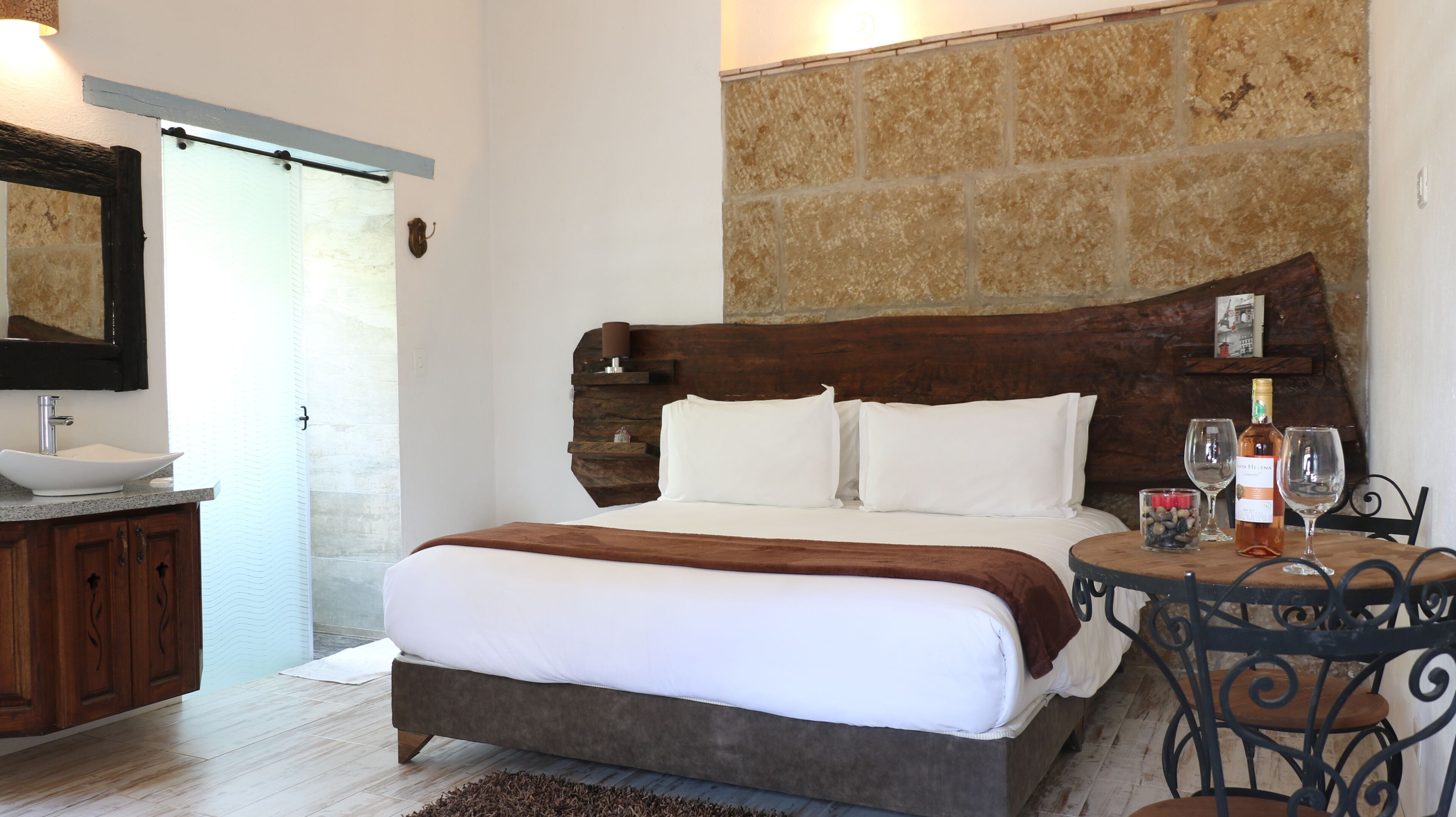 A bedroom with a wooden headboard, a bed with white bedding, a nightstand with a bottle and glasses, and a bathroom with a sink and mirror.