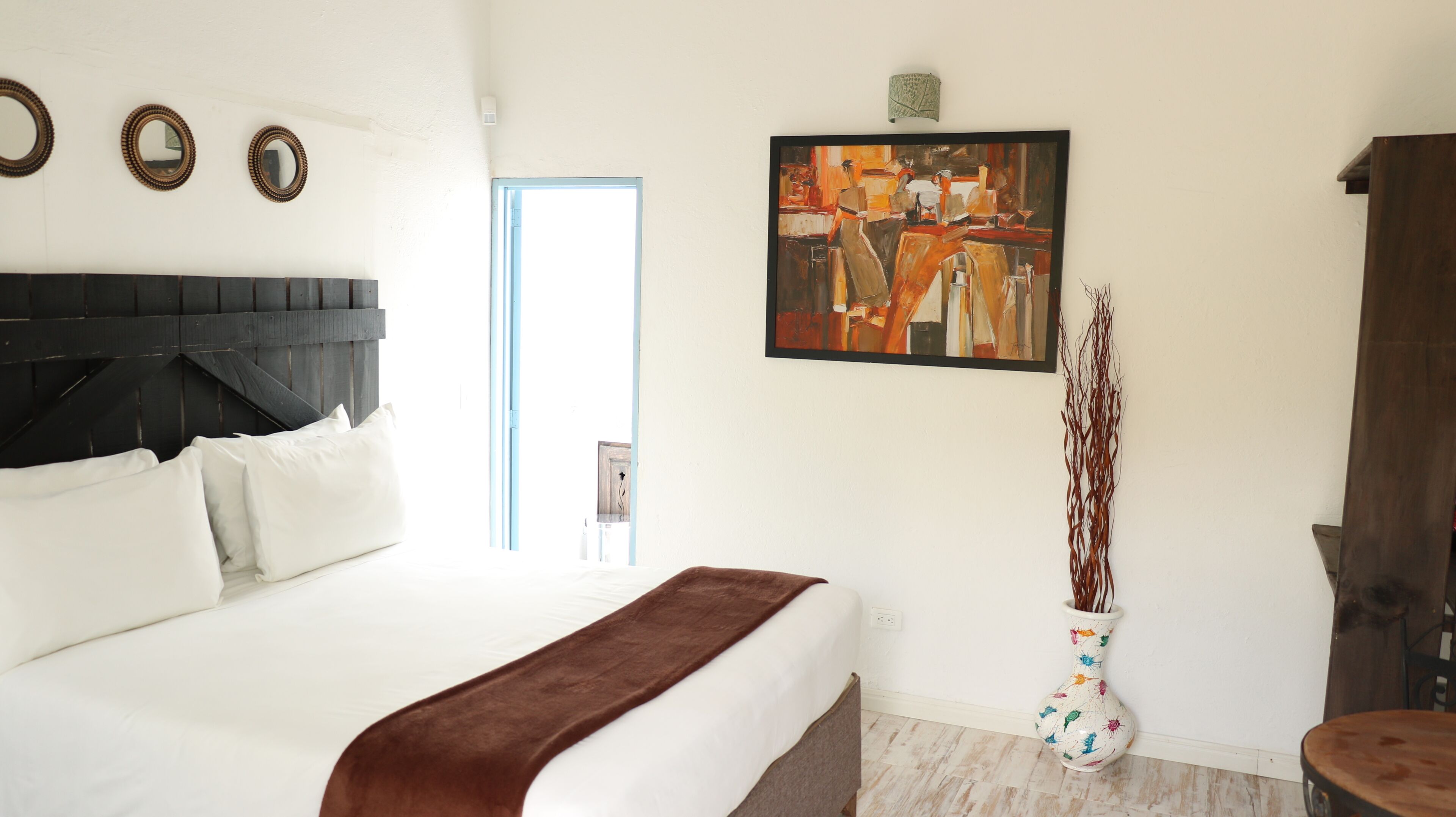 A hotel room with a bed, a painting, a vase with branches, and a wooden headboard.