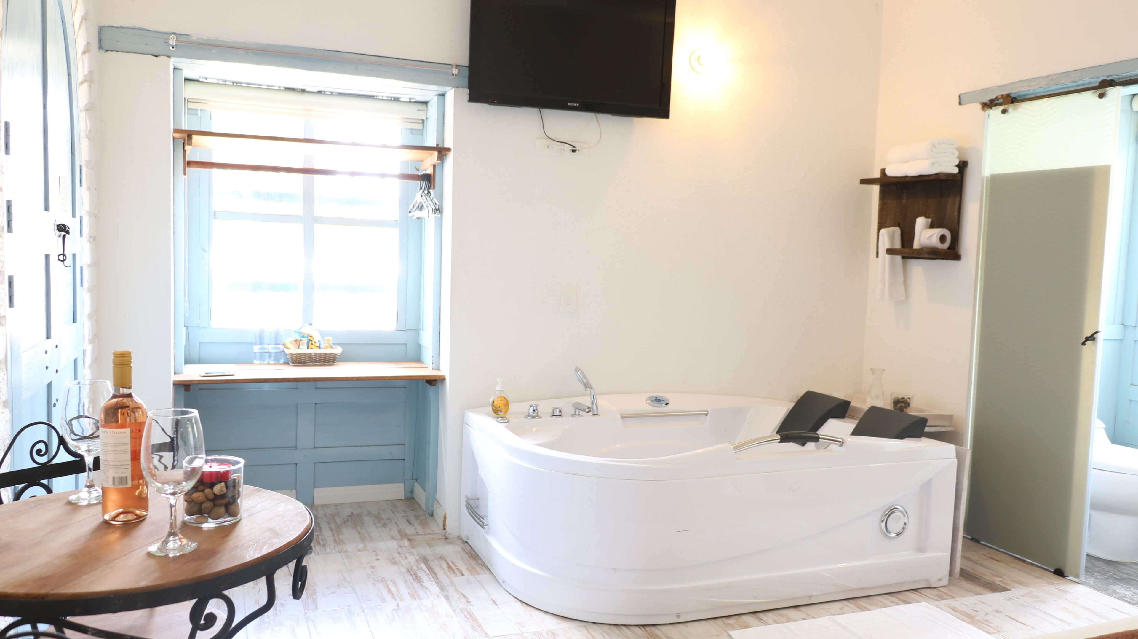Habitación con una bañera blanca, una mesa de madera con copas y botellas de vino, una ventana con vistas y un televisor de pantalla plana montado en la pared.