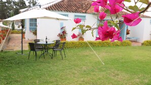 A patio with a table and chairs, a white building with blue windows, and a garden with pink flowers. - Hotel Boutique Santa Helena (Villa de Leyva)