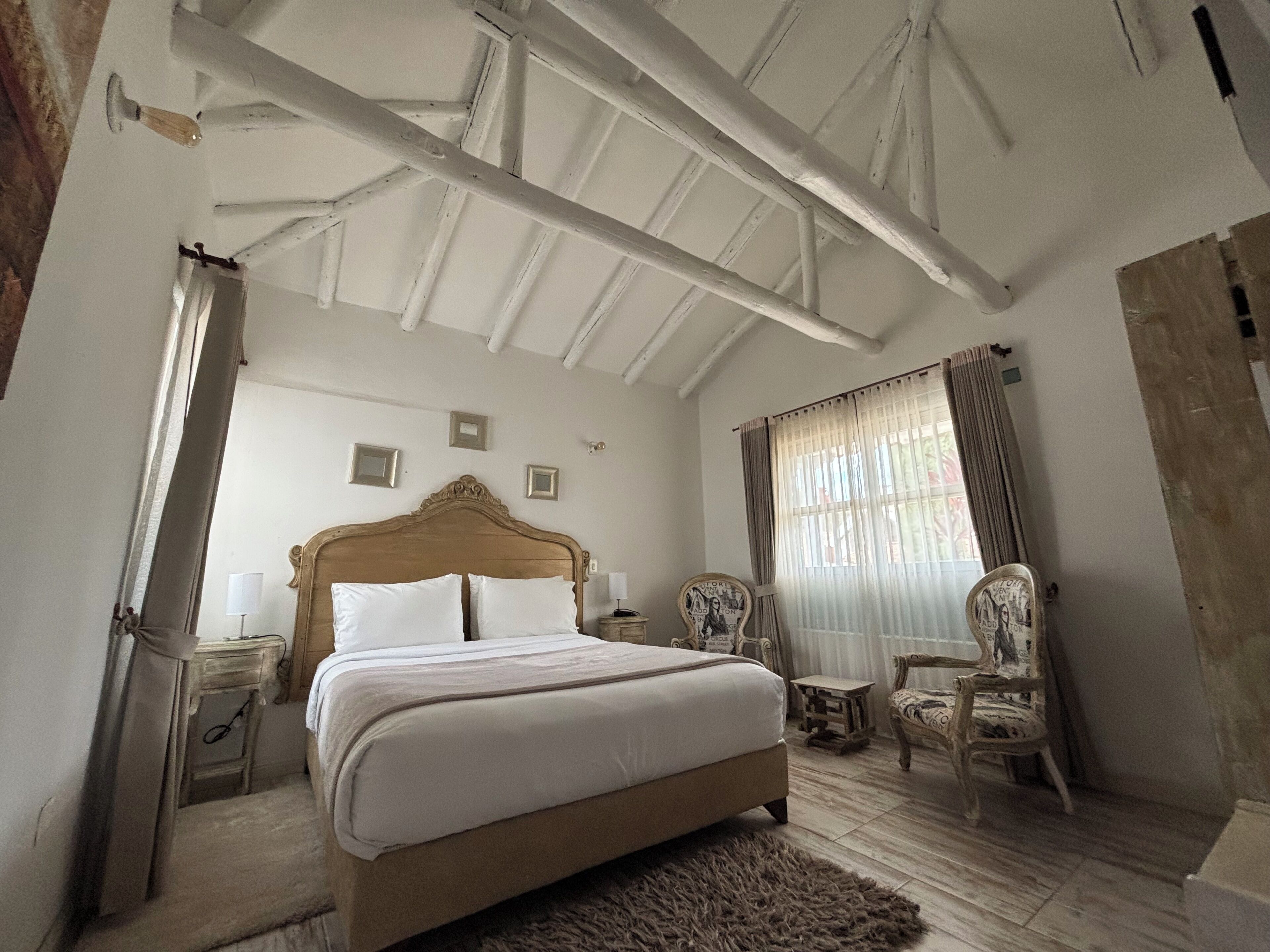 A bedroom with a large bed, a wooden headboard, a nightstand, a chair, and a window with curtains.