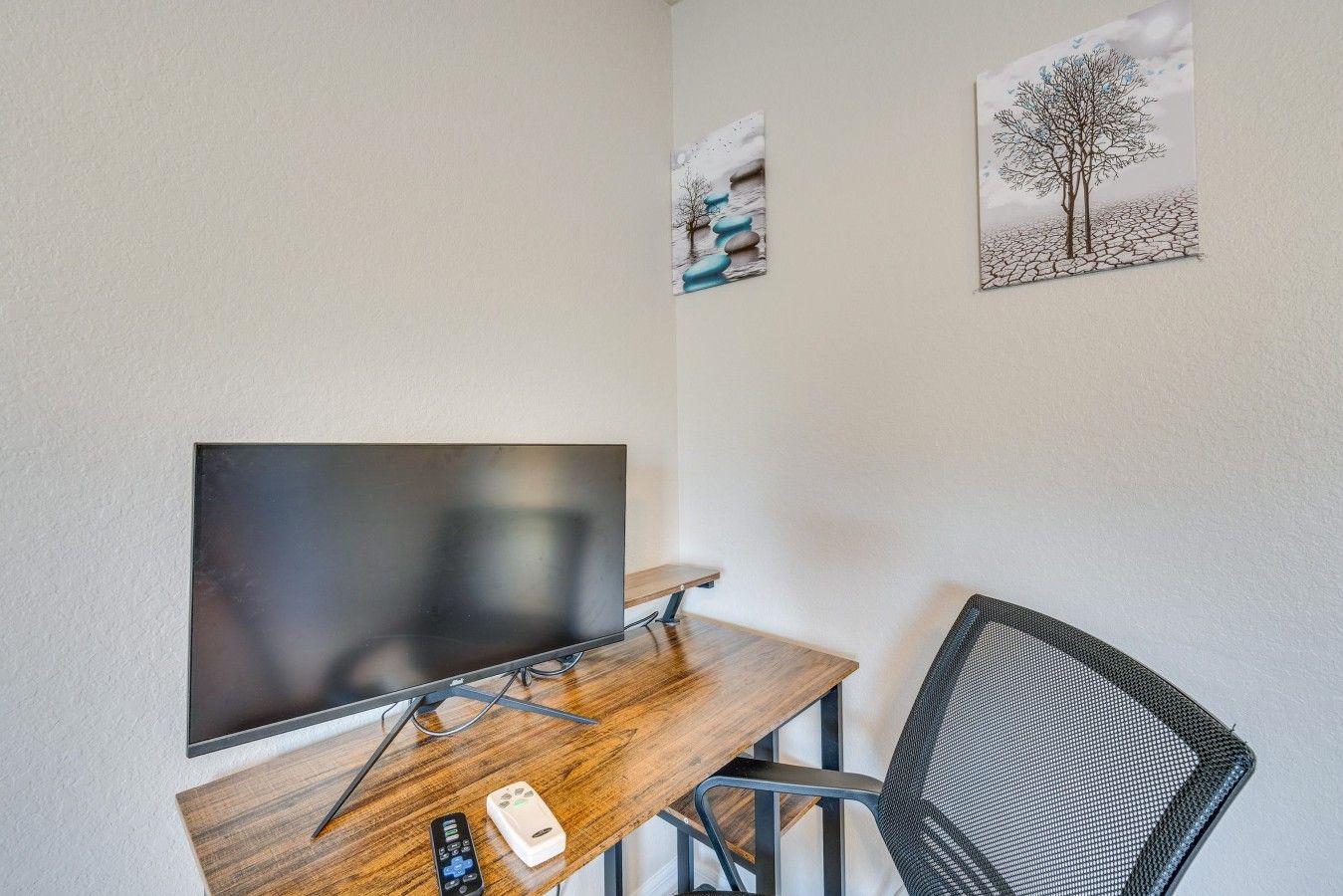 A room with a flat-screen TV mounted on the wall, a wooden desk with a chair, a remote control, and a small device on the desk.