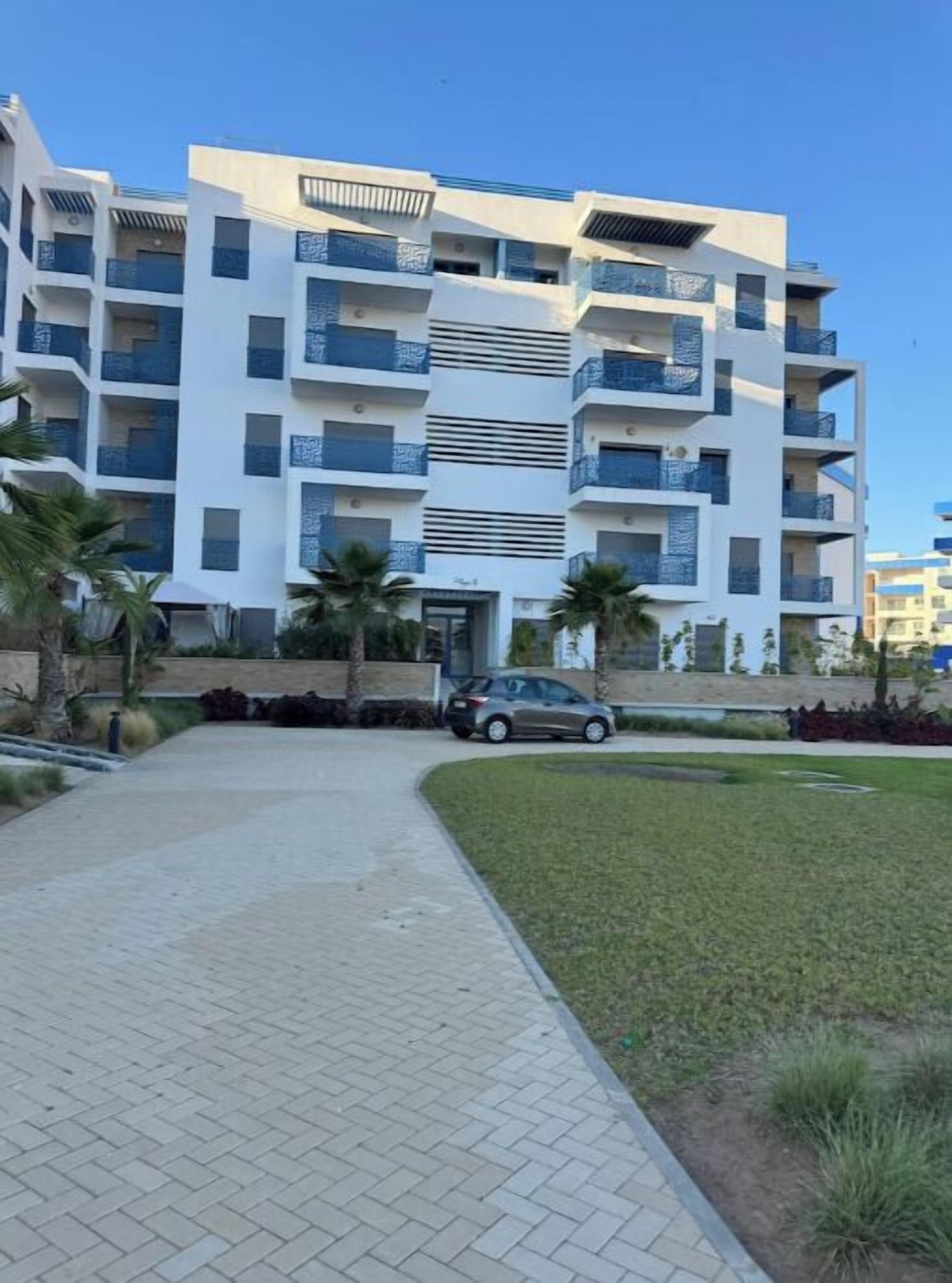 A multi-story residential building with balconies, a paved walkway, and a car parked in front.