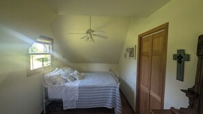 Ein Schlafzimmer mit einem Einzelbett, einem Fenster, einem Deckenventilator, einer Holztür und einem Kreuz an der Wand.