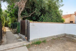 Un chemin menant à un mur blanc percé d’une porte, le tout entouré de verdure et d’une clôture.