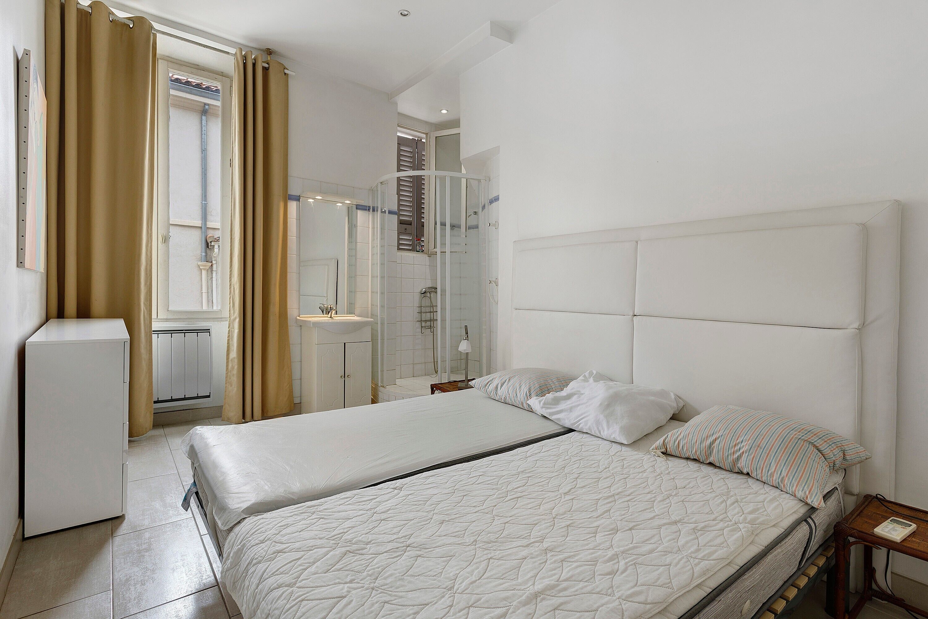 A hotel room with a bed, a bedside table, a mirror, a bathroom with a shower, and a window with curtains.