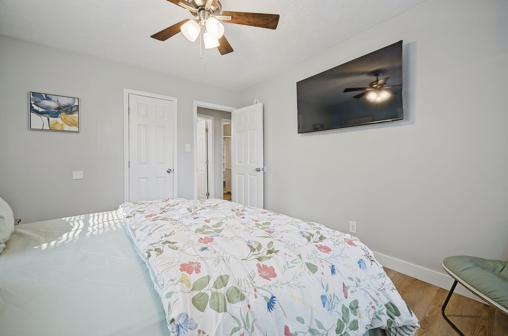 A bedroom with a bed, a ceiling fan, a door, and a wall-mounted TV.