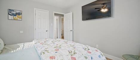 A bedroom with a bed, a ceiling fan, a door, and a wall-mounted TV.