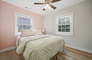 Un dormitorio con una cama, dos ventanas, un ventilador de techo y piso de madera.