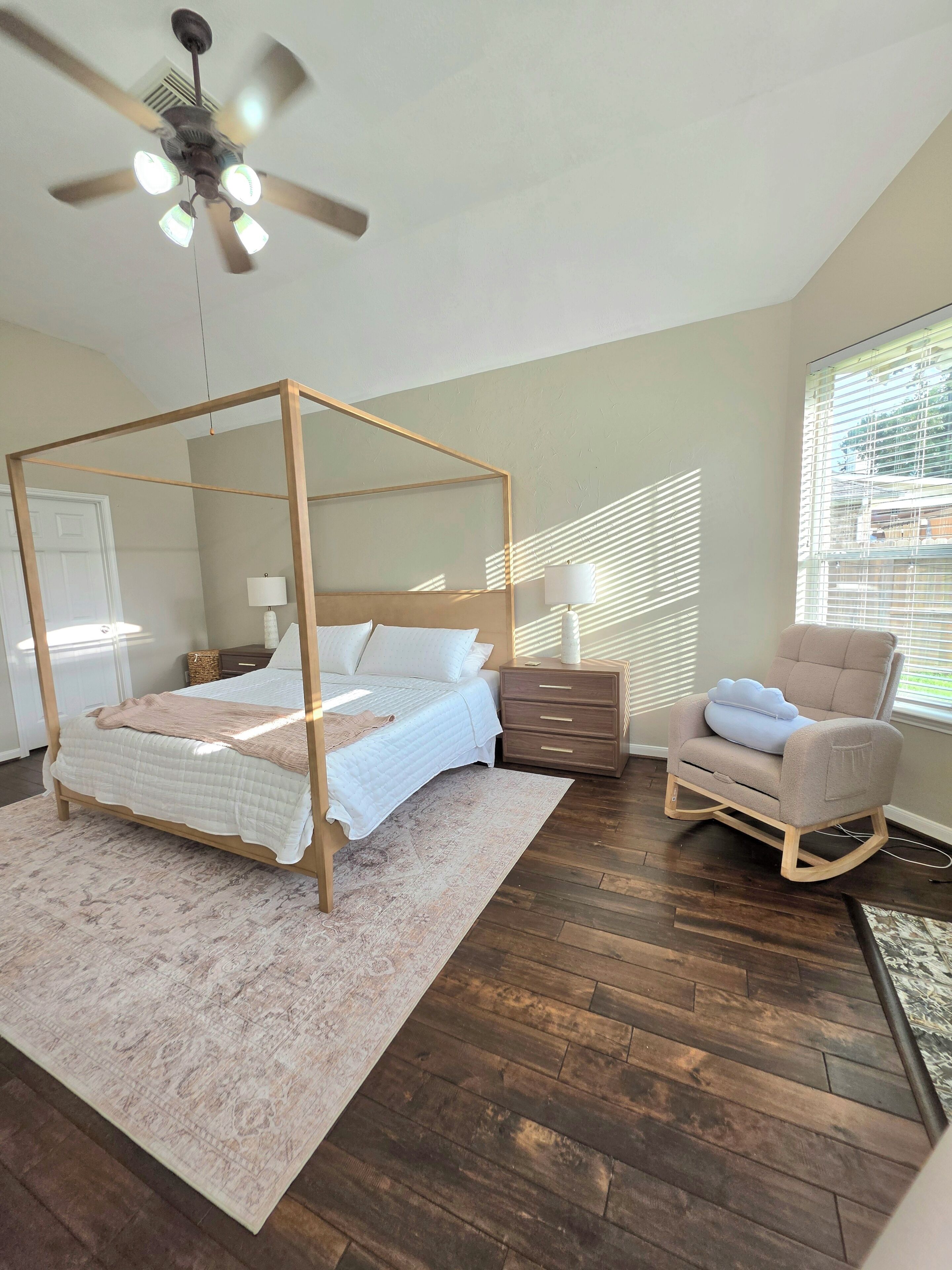 A bedroom with a four-poster bed, a rocking chair, a bedside table, and a ceiling fan.