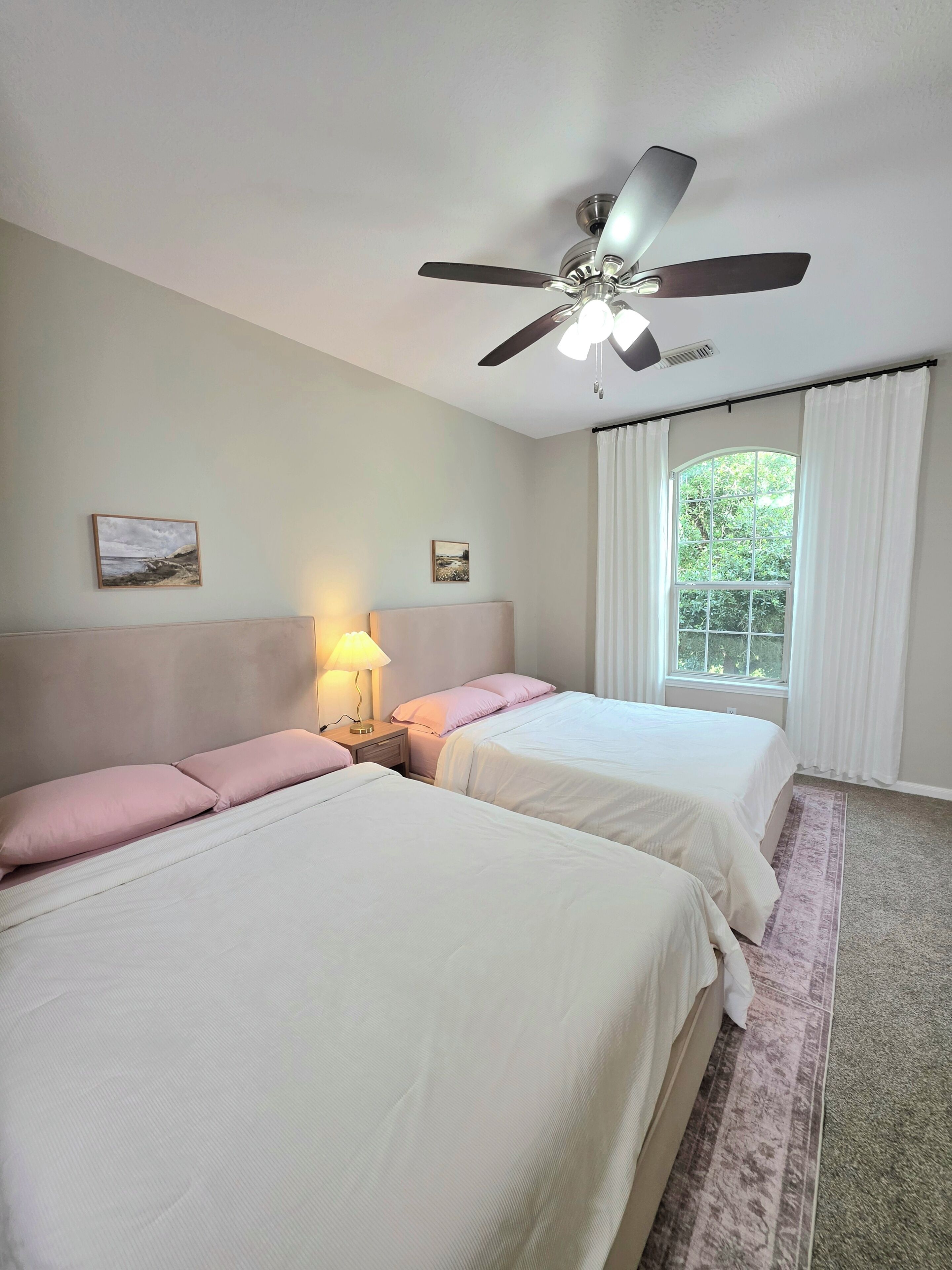 A hotel room with two beds, a ceiling fan, a window with curtains, and a nightstand with a lamp.