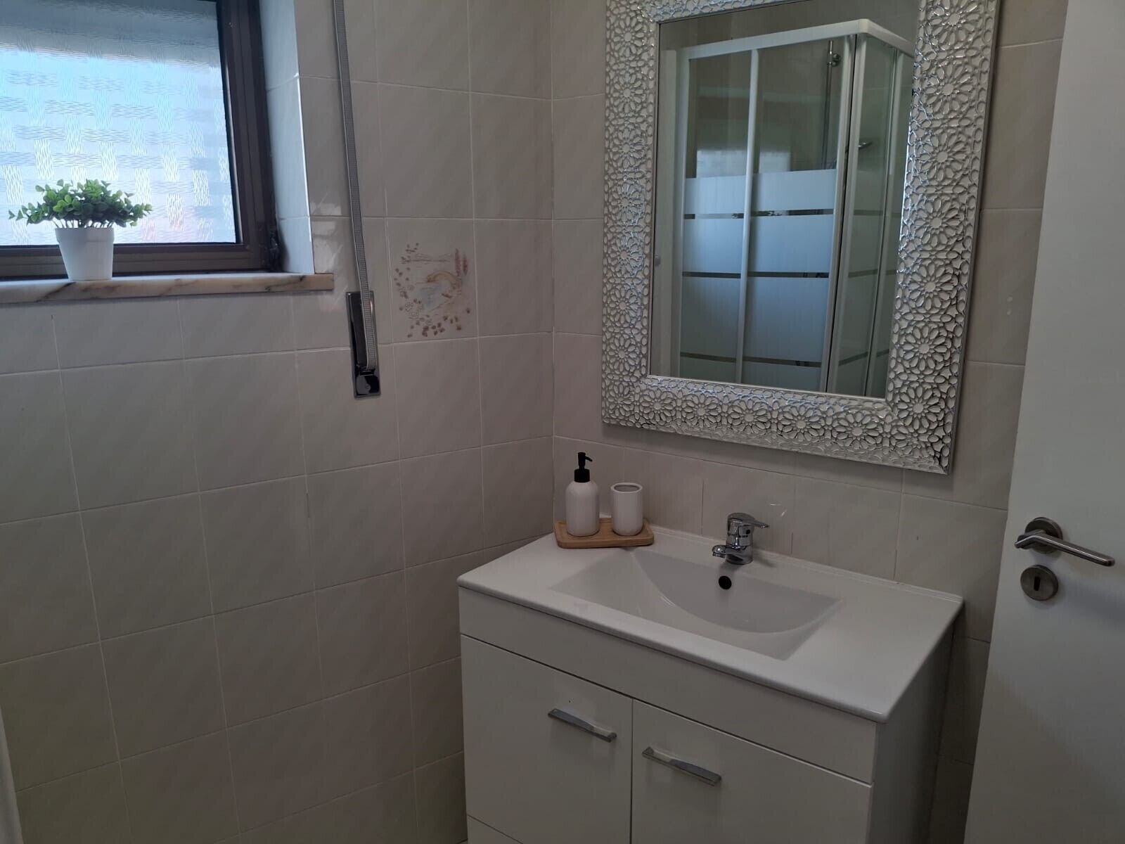 Une salle de bain avec un lavabo blanc, un miroir orné d’un cadre floral et une petite fenêtre avec un pot de fleurs.