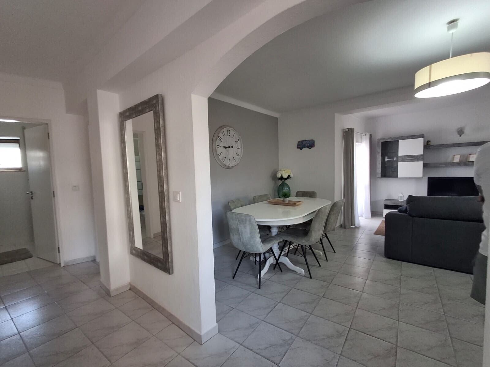 A modern living room with a dining area, grey tiled flooring, a large mirror, a clock, and a TV.
