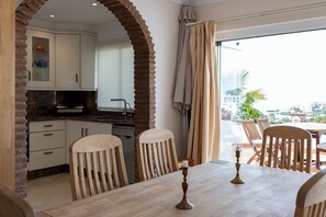 A dining area with a wooden table and chairs, a kitchen with white cabinets, and a view through a glass door. - La Pomonita - Two Bedroom Apartment, Sleeps 4 (Nerja)