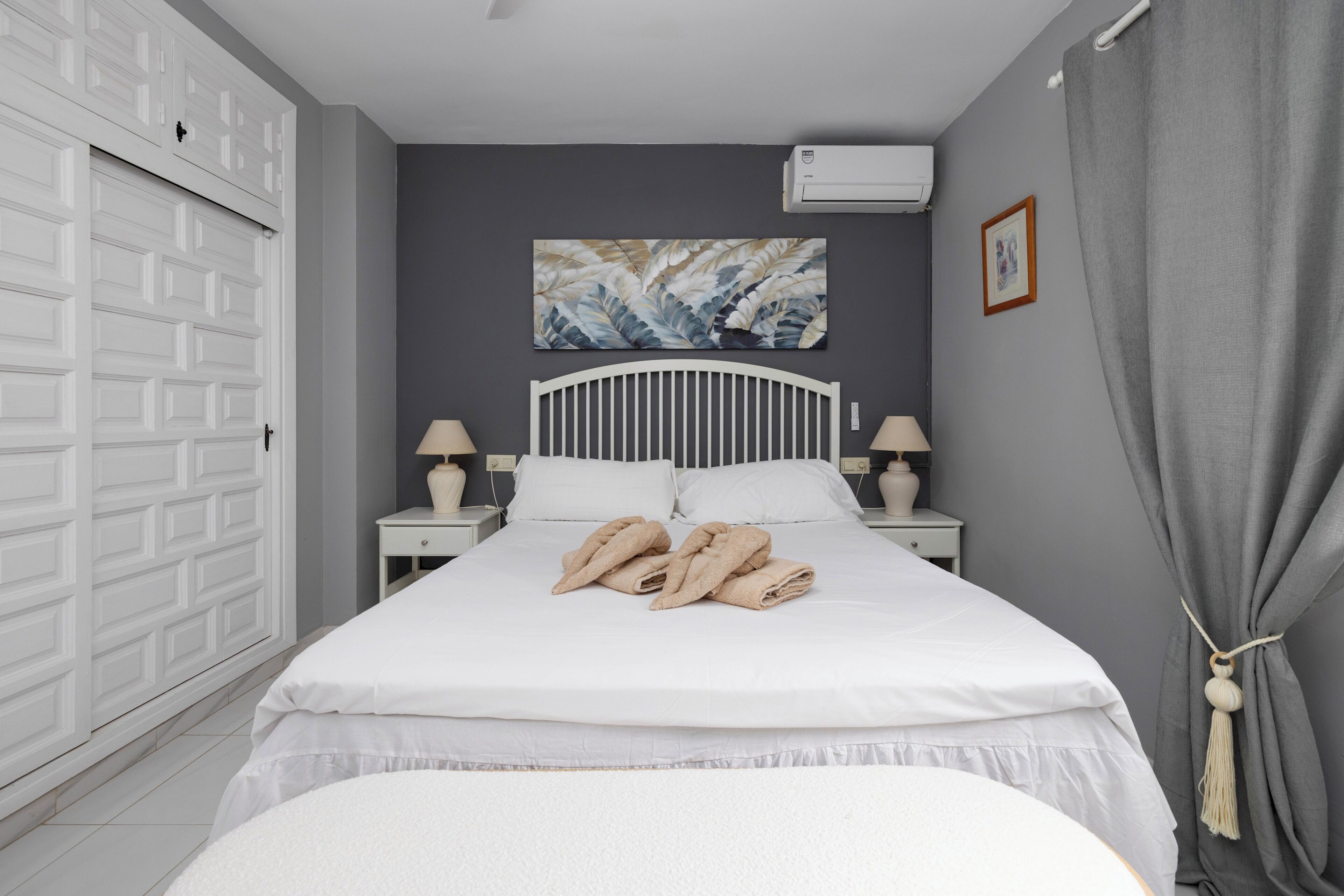 A bedroom with a bed, two bedside tables, a wall-mounted air conditioner, a painting, and a door.