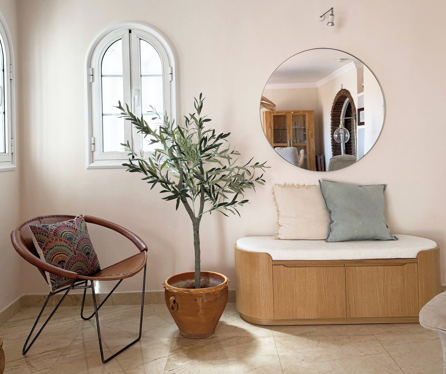 Une pièce comprenant un banc en bois, une plante en pot, une chaise avec un coussin à motifs et un miroir rond accroché au mur.