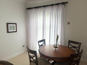 Uma sala de jantar com uma mesa redonda de madeira, quatro cadeiras, um quadro na parede e uma janela com cortinas brancas.