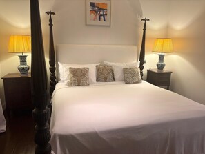 Ein Bett mit Baldachin, ausgestattet mit weißen Leinentüchern und gemusterten Kissen, steht zwischen zwei Nachttischen mit Lampen, in einem Zimmer mit schlichter Wand und einem gerahmten Bild über dem Bett.