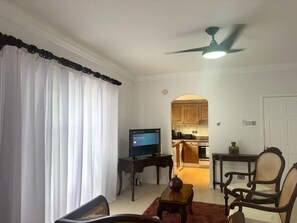 Ein Wohnzimmer mit Fernseher, Couchtisch und Sessel. Durch einen Durchgang ist eine Küche zu sehen.