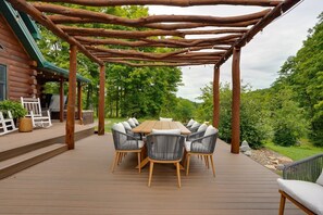 Een houten terras met een pergola, een ronde houten tafel en zes stoelen met kussens.